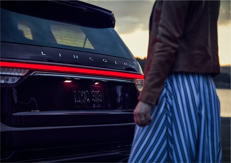 A person is shown near the rear of a 2023 Lincoln Aviator® SUV as the Lincoln Embrace illuminates the rear lights | Sentry Lincoln in Medford MA