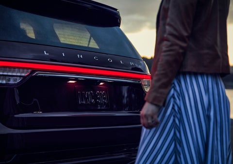 A person is shown near the rear of a 2024 Lincoln Aviator® SUV as the Lincoln Embrace illuminates the rear lights | Sentry Lincoln in Medford MA
